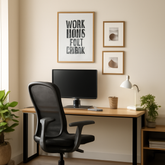 A minimalist home office with natural light streaming in through a large window. The setup includes a wooden desk with a black ergonomic chair, a computer monitor, a desk lamp, and a small potted plant.